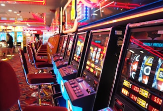 A brightly lit casino interior featuring a row of colorful slot machines. The neon lights create a vibrant atmosphere, with patterned carpeting adding to the decor. Stools are aligned neatly in front of each machine.